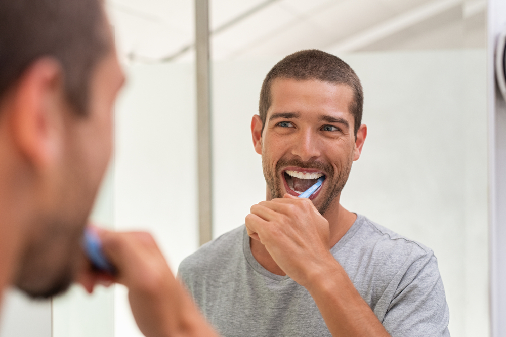 Smiling young man with toothbrush cleaning teeth and looking mirror in the bathroom