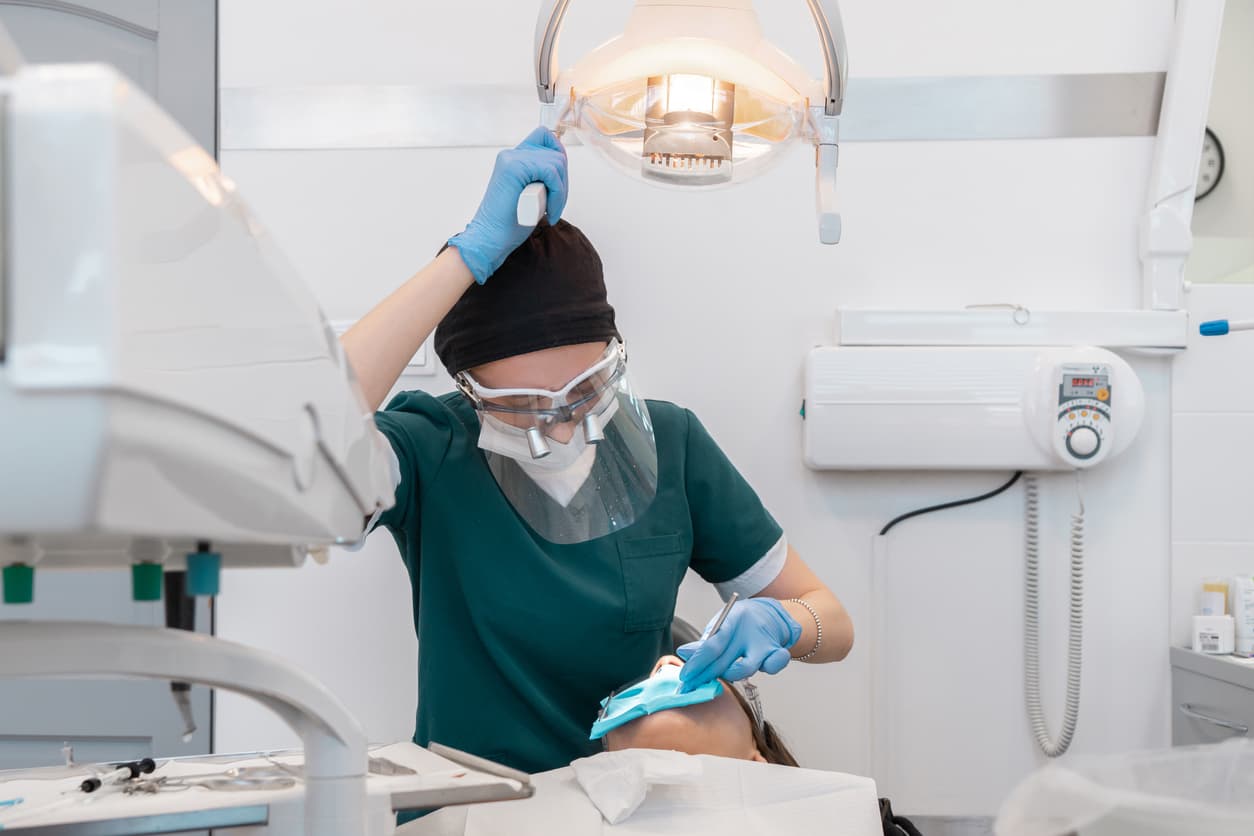A dentist wearing protective equipment examines a patient under an overhead dental light. The image highlights safe dental care, routine examinations, infection control, and a modern clinical environment.