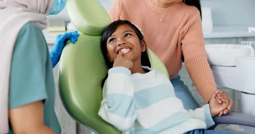 Young girl smiling during a pediatric dental checkup with her parent nearby