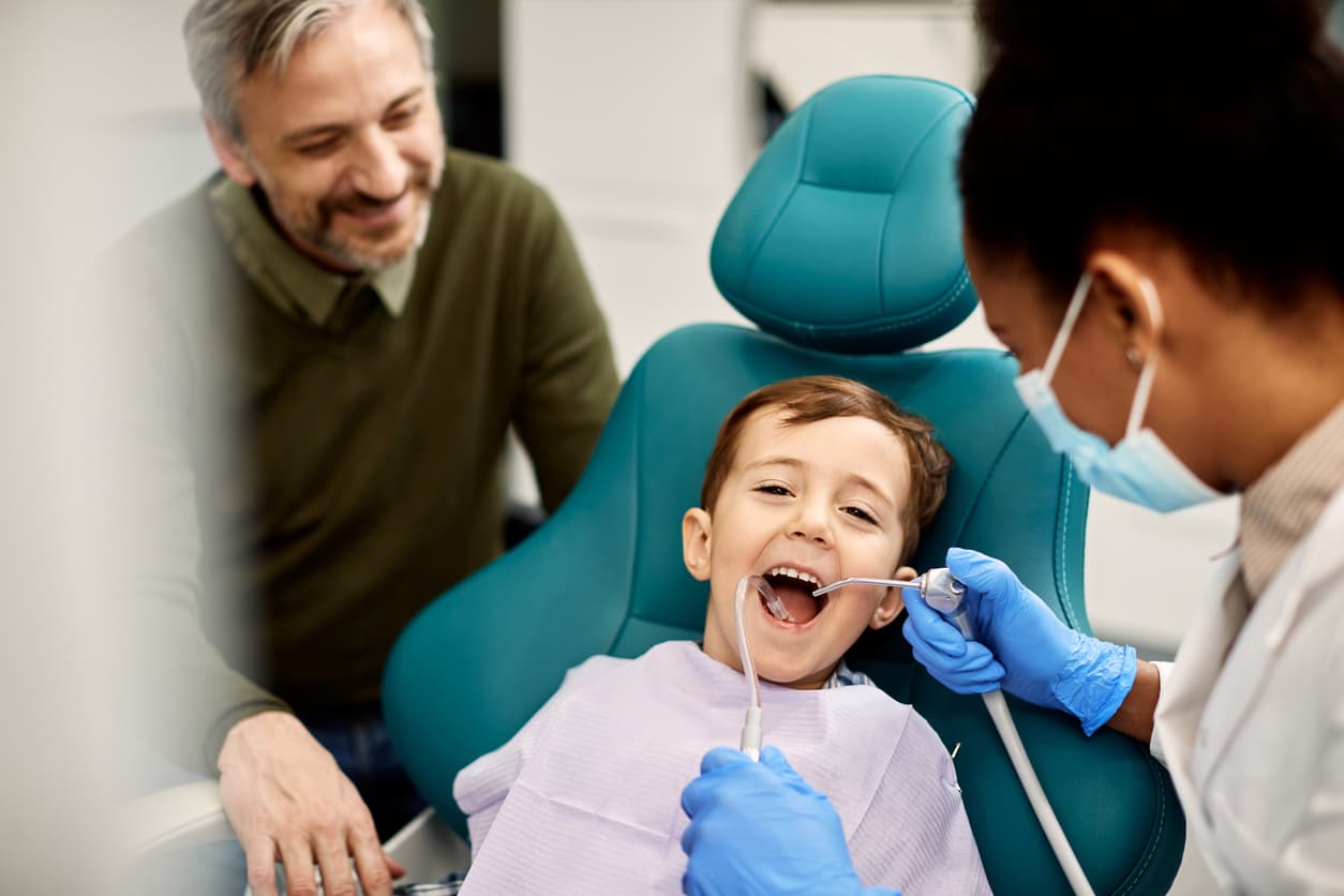 A young child smiles during a dental examination while a dentist performs a gentle checkup and a parent looks on. The image highlights family dentistry, pediatric dental care, and a positive experience for children.