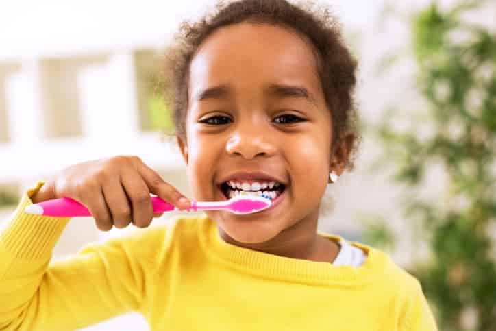 Smiling young girl brushing her teeth with a pink toothbrush at home
