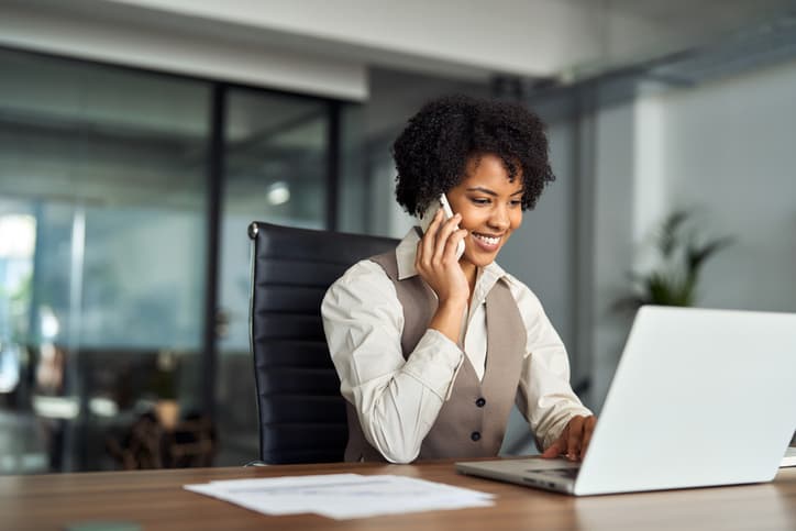 Businesswoman smiling while talking on the phone and working on a laptop at her desk”