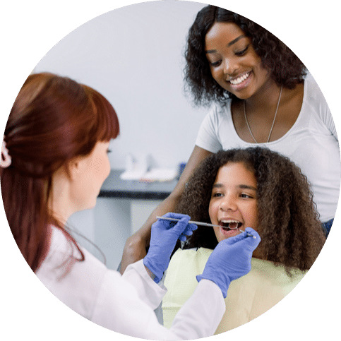 Dentist examining a young girl’s teeth while her mother stands beside her