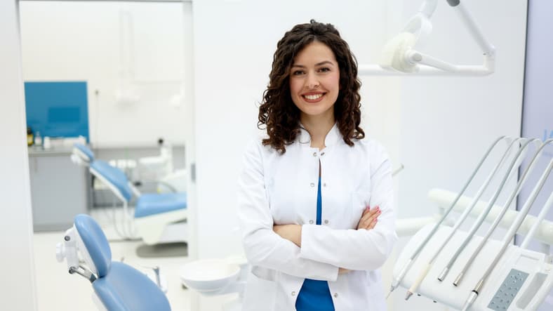 Female dentist standing in bright dental clinic with equipment, smiling with arms crossed