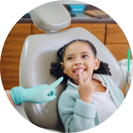 Young girl pointing at her teeth while sitting in a dental chair during a pediatric dental visit”
