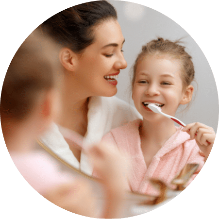Mother helping her daughter brush her teeth in front of a bathroom mirror