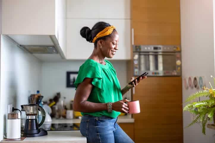 Woman holding a mug and checking her phone while standing in a kitchen