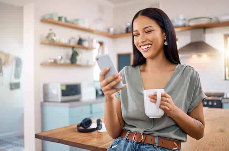 Woman smiling while checking her phone and holding a mug in a kitchen