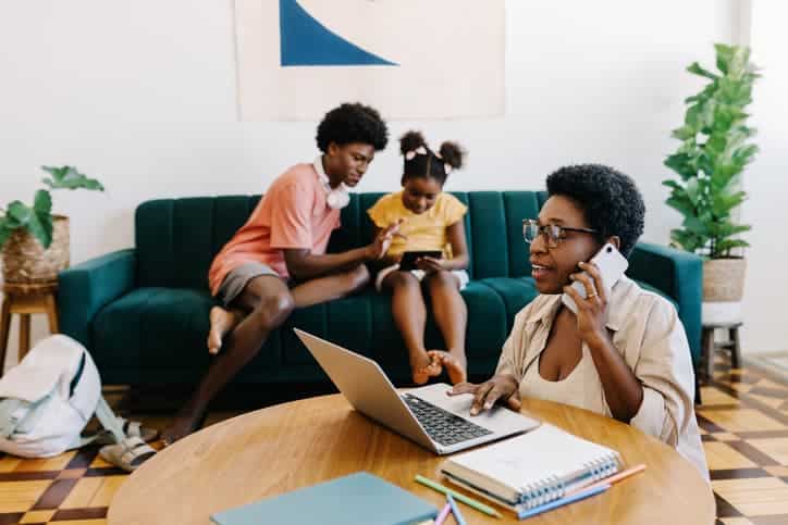 Woman talking on the phone while working on a laptop as children sit on a couch behind her