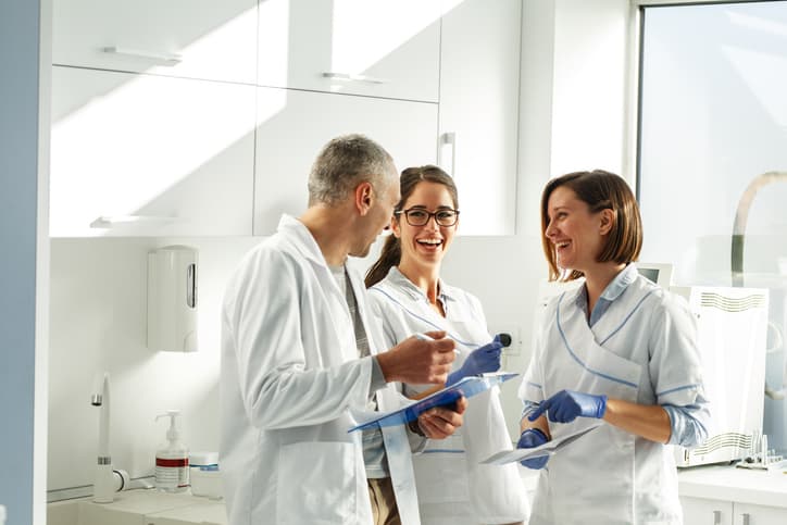 Three dental professionals review notes and discuss patient care in a bright dental clinic. The image represents teamwork, collaboration, treatment planning, and a coordinated approach to providing high-quality dental care.