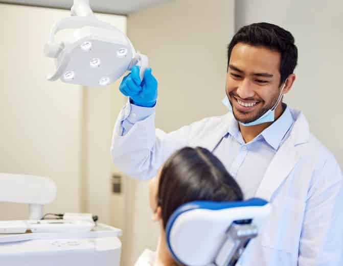 A smiling dentist adjusts the overhead examination light while greeting a patient in the dental chair. The image conveys a welcoming atmosphere, compassionate care, and a positive dental experience.