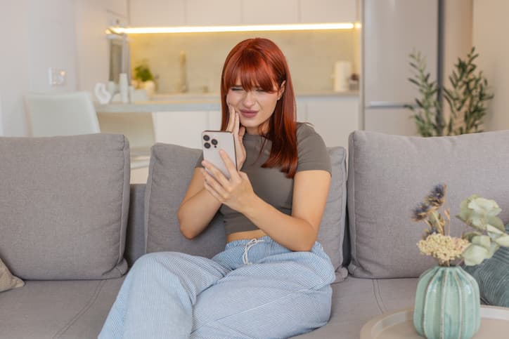 Person at home holding their jaw and looking at a smartphone while experiencing severe tooth pain, illustrating the discomfort of a dental emergency before seeking professional treatment.