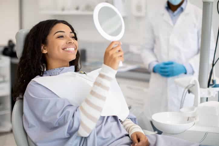 Smiling dental patient looking at her teeth in a handheld mirror after receiving dental care while seated in a dental chair