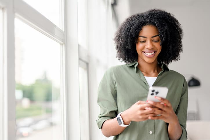 Smiling woman using her smartphone while standing by a bright window, showing a healthy, confident smile